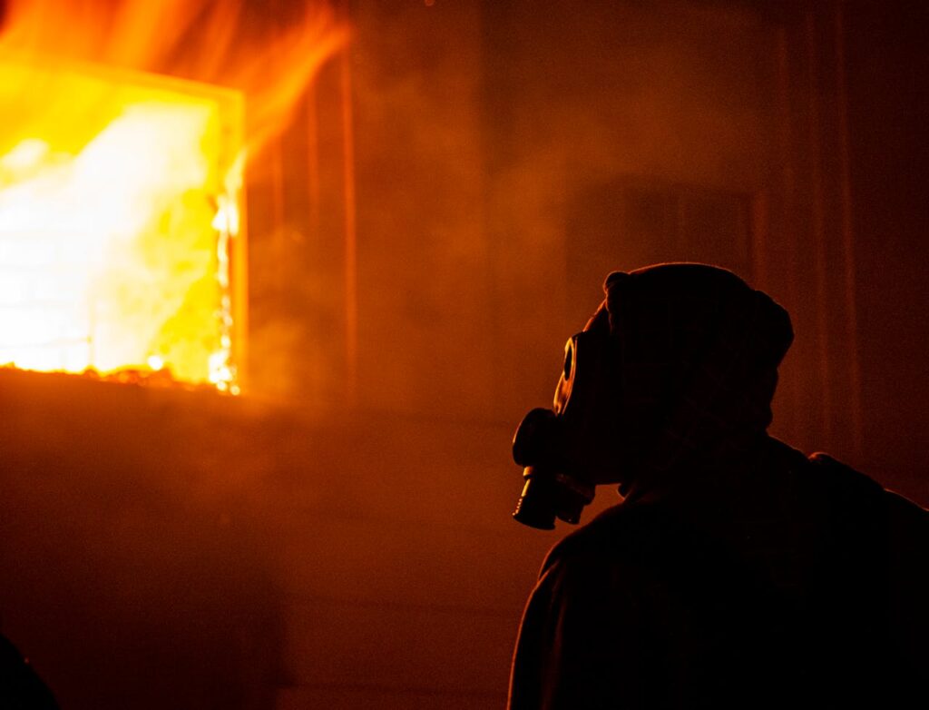 Silhouette of a person wearing a gas mask in front of a burning building at night.
